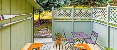 En träveranda med ett bord och stolar, omgiven av ett grönt staket och tät vegetation.
