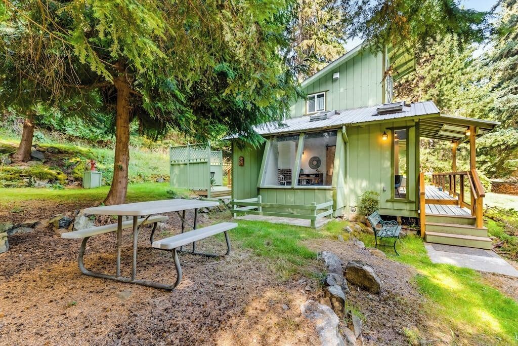 A green wooden house with a porch, a picnic table, and a grill area.