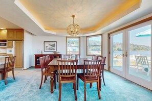 A dining area with a large table and chairs, a chandelier, and a view of the outdoors through large windows. - Charming Waterfront Home in Greenbank with Direct Beach Access and Stunning Ocean Views (Greenbank)
