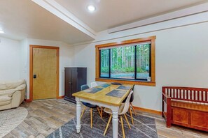 Uma sala de jantar com mesa, cadeiras, um banco, uma porta e uma janela com vista para árvores.