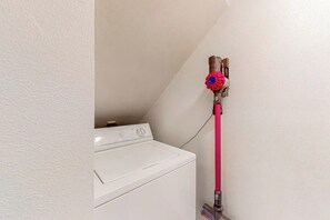 Máquina de lavar roupa branca num canto, com uma mangueira rosa ligada a uma torneira fixada na parede.