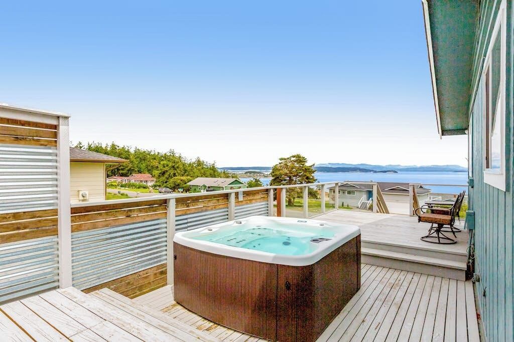 Un jacuzzi en una terraza de madera con vista al océano y casas al fondo.