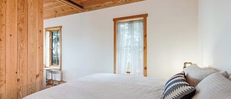A bedroom with a large bed, two windows, wooden ceiling beams, and a small desk.