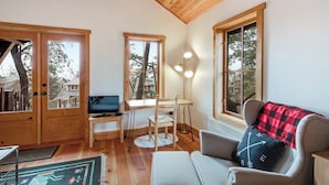 Ein gemütliches Zimmer mit Holzbalkendecke, einem Schreibtisch mit Computer, einem Sessel, einer Couch und einem großen Fenster.
