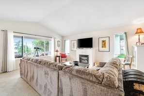 A living room with a large sectional sofa, a fireplace, a television, and a view through the glass doors. - Charming Island House with Beach Access, Tennis Courts, and Backyard (Coupeville)