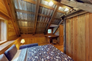Una stanza con soffitto spiovente, pareti in legno, un letto con copriletto floreale, una scrivania e una piccola sedia.