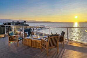 Un terrazzo in legno con posti a sedere all'aperto, un tavolo da pranzo apparecchiato con piatti e bicchieri e vista sul tramonto sull'acqua.