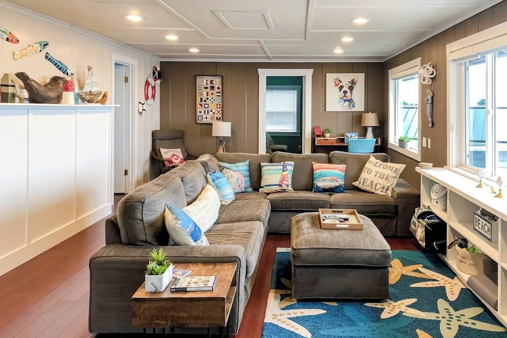 Una sala de estar con sofá seccional, reposapiés y una mesa de centro. Hay una estantería con un cartel que dice 'BEACH', una ventana con persianas y una puerta que da a otra habitación.