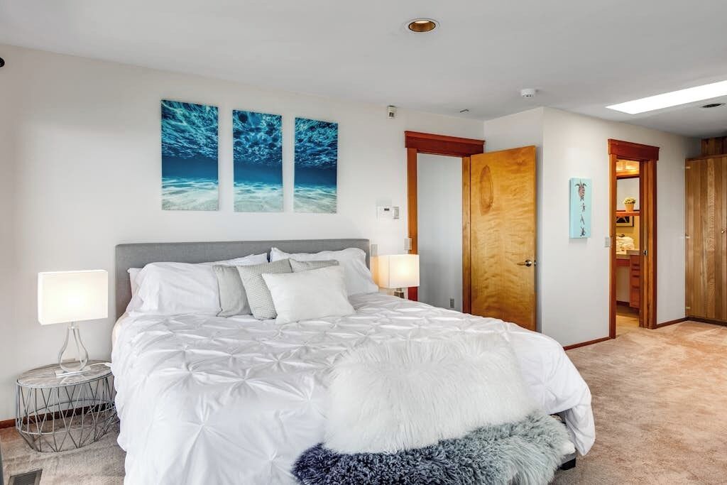 A bedroom with a large bed, two wall-mounted paintings, a wooden door, and a small round table with a lamp.