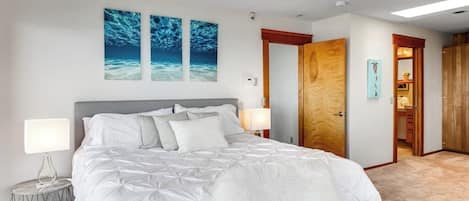 A bedroom with a large bed, two wall-mounted paintings, a wooden door, and a small round table with a lamp.