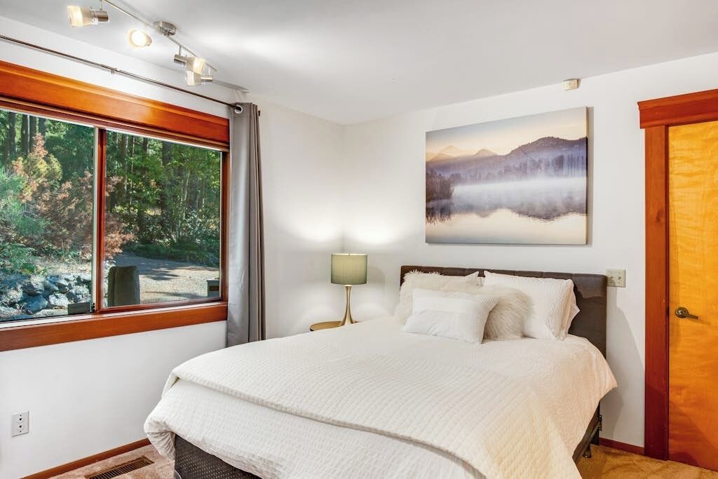 Un dormitorio con una cama grande, una ventana con vistas a árboles, un cuadro en la pared y una lámpara.