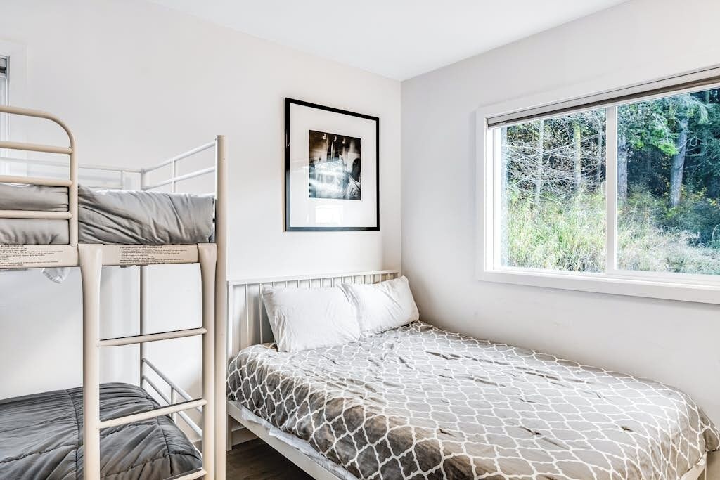 A bunk bed with a white headboard and a single bed with a patterned bedspread. There is a framed picture on the wall and a window with a view of trees.