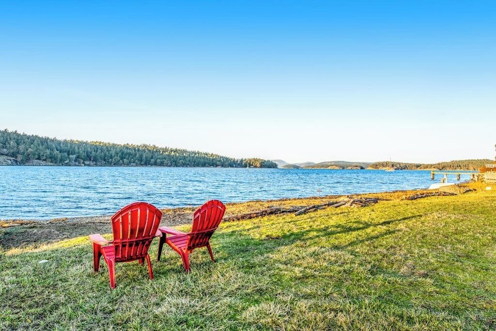 Two red chairs by a lake with a grassy area and a forested hill in the background.