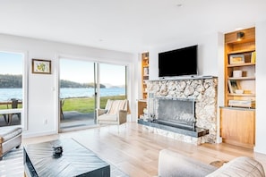 A modern living room with a stone fireplace, a flat-screen TV, and a view of the lake. - Accessible Waterfront Holiday Home with Bay Views and Family Friendly Features (Lopez Island)