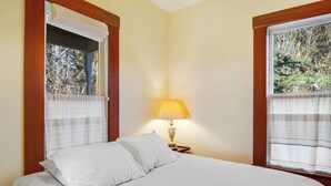 A bedroom with a bed, a nightstand with a lamp, and windows with curtains. - Classic farmhouse-style home with front porch & amazing sunset views - walk to town (Lopez Island)