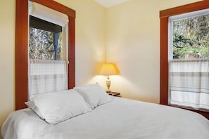 A bedroom with a bed, a nightstand with a lamp, and windows with curtains. - Classic farmhouse-style home with front porch & amazing sunset views - walk to town (Lopez Island)