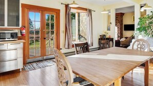 A dining area with a wooden table and chairs, a kitchen with a microwave, and a large window with curtains. - Classic farmhouse-style home with front porch & amazing sunset views - walk to town (Lopez Island)
