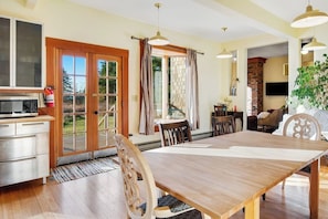 A dining area with a wooden table and chairs, a kitchen with a microwave, and a large window with curtains. - Classic farmhouse-style home with front porch & amazing sunset views - walk to town (Lopez Island)