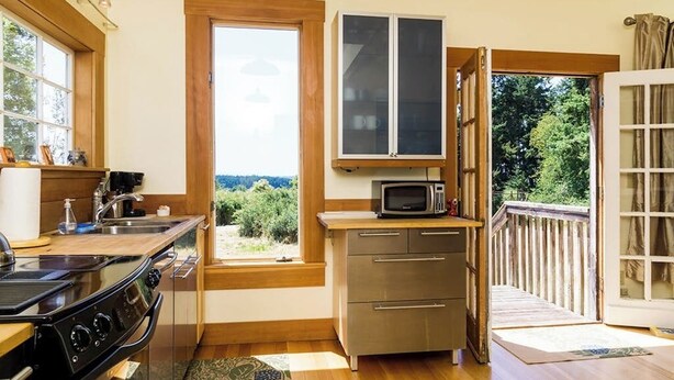 A compact kitchen with a stove, microwave, and cabinets, a large window with a view, and a wooden deck outside. - Classic farmhouse-style home with front porch & amazing sunset views - walk to town (Lopez Island)