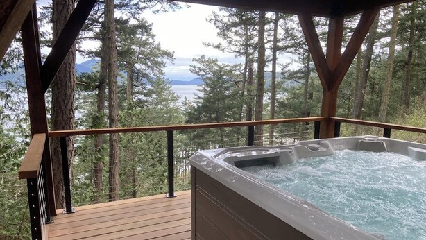 A wooden deck with a hot tub and a view of a forest and water. - Renovated 3 Bedroom Eastsound Home with Hot Tub Fire Pit and Stunning Sound Views (Eastsound)