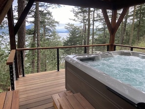 A wooden deck with a hot tub and a view of a forest and water. - Renovated 3 Bedroom Eastsound Home with Hot Tub Fire Pit and Stunning Sound Views (Eastsound)