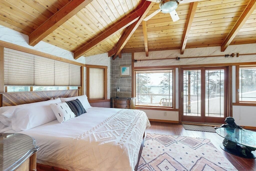 A bedroom with a large bed, wooden ceiling beams, a glass table, and a view of the outdoors.