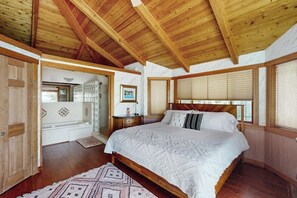 A bedroom with a wooden ceiling, a large bed, a window with blinds, and a bathroom with a glass shower enclosure. - Pet Friendly Waterfront Escape with Fire Pit, Hot Tub and Mountain Views on Lopez Island (Lopez Island)