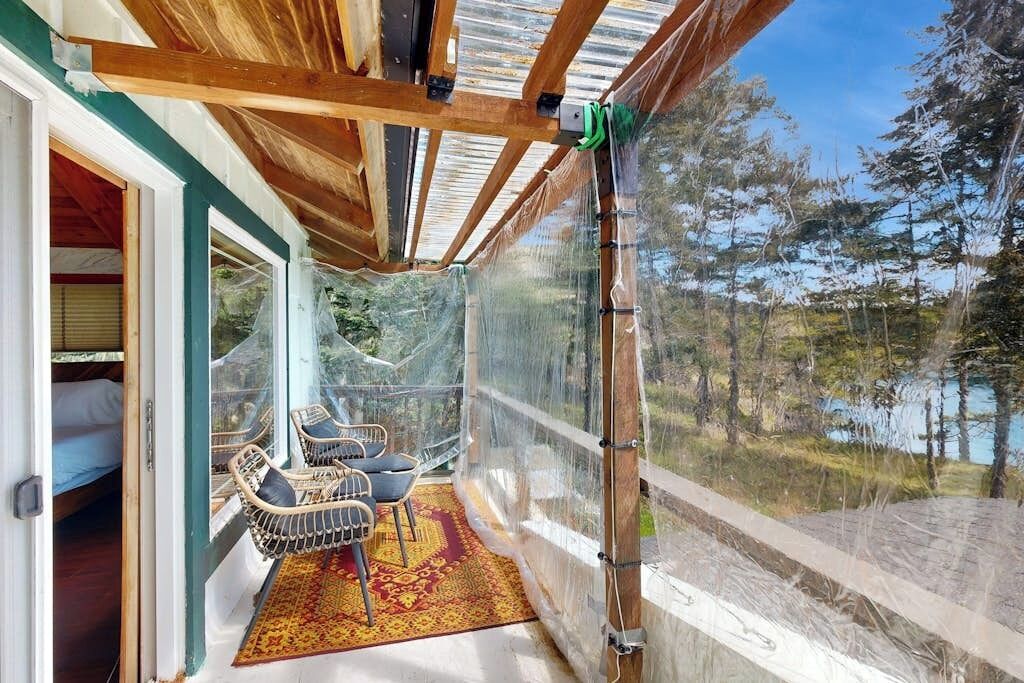 En overdækket balkon med udsigt over skov og sø. Balkonen har fletstole og en tæppe med et mønster.