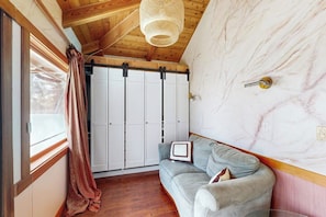 A room with a wooden ceiling, a white wall with a marble-like pattern, a sliding door with white panels, a grey sofa with cushions, and a window with a curtain. - Pet Friendly Waterfront Escape with Fire Pit, Hot Tub and Mountain Views on Lopez Island (Lopez Island)