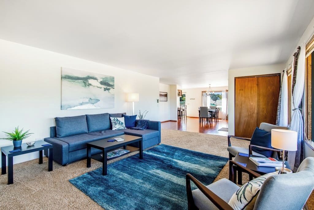 Un salon moderne avec un canapé, une table basse et un tapis. On aperçoit un coin repas au fond de la pièce.
