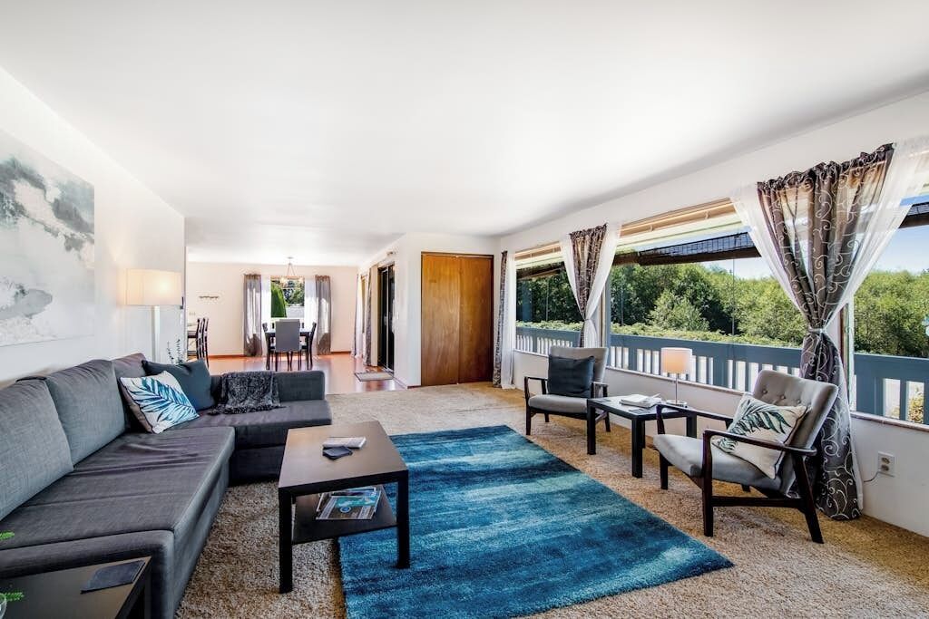 A modern living room with a sofa, coffee table, and chairs. There is a balcony with curtains and a view of greenery.