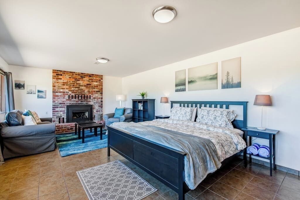 A bedroom with a brick fireplace, a bed with a blue headboard, a grey sofa, and a small table.