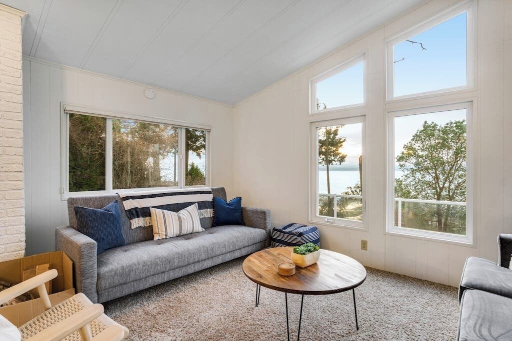 A room with a grey sofa, a round wooden coffee table, and large windows offering a view of trees and a body of water.