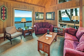 A cozy room with a wooden interior, a large window with a view of the sea, a purple sofa, a striped chair, and a wooden coffee table.