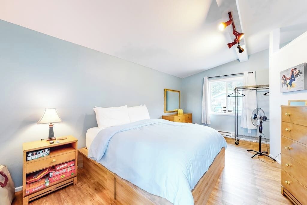 A bedroom with a bed, bedside table, lamp, dresser, and a window with a view.