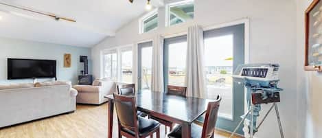 A modern living room with a flat-screen TV, a sofa, a dining table with chairs, and a large window with curtains.