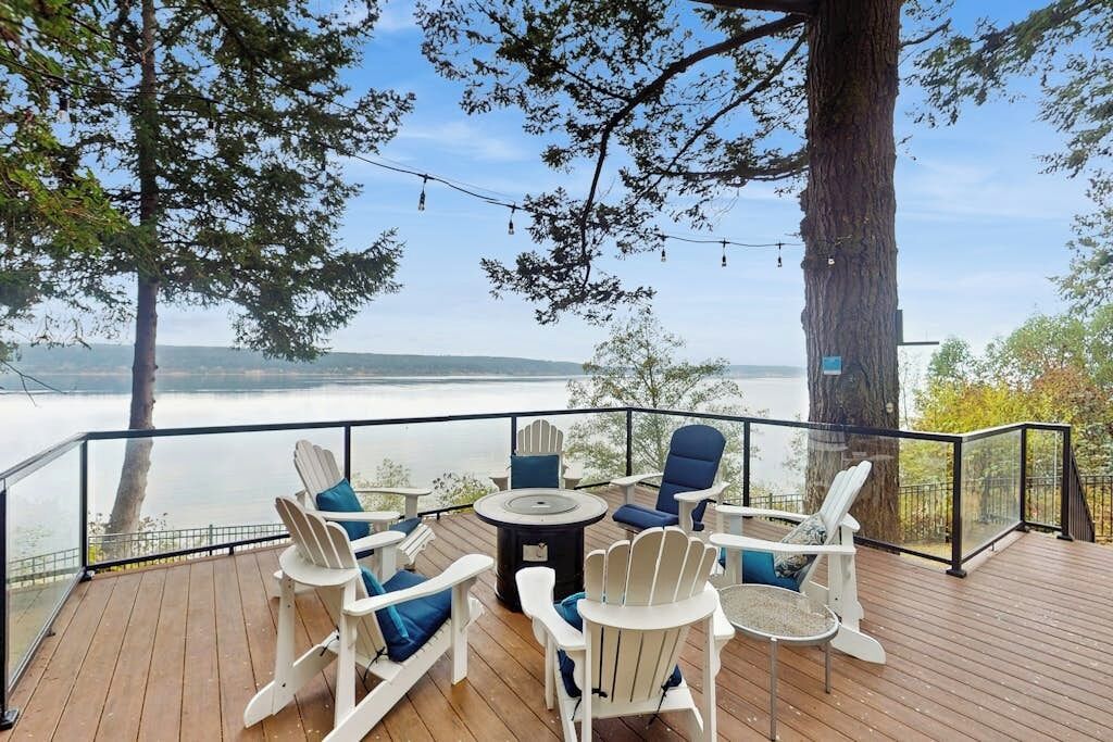 A deck with Adirondack chairs and a round table, overlooking a body of water.