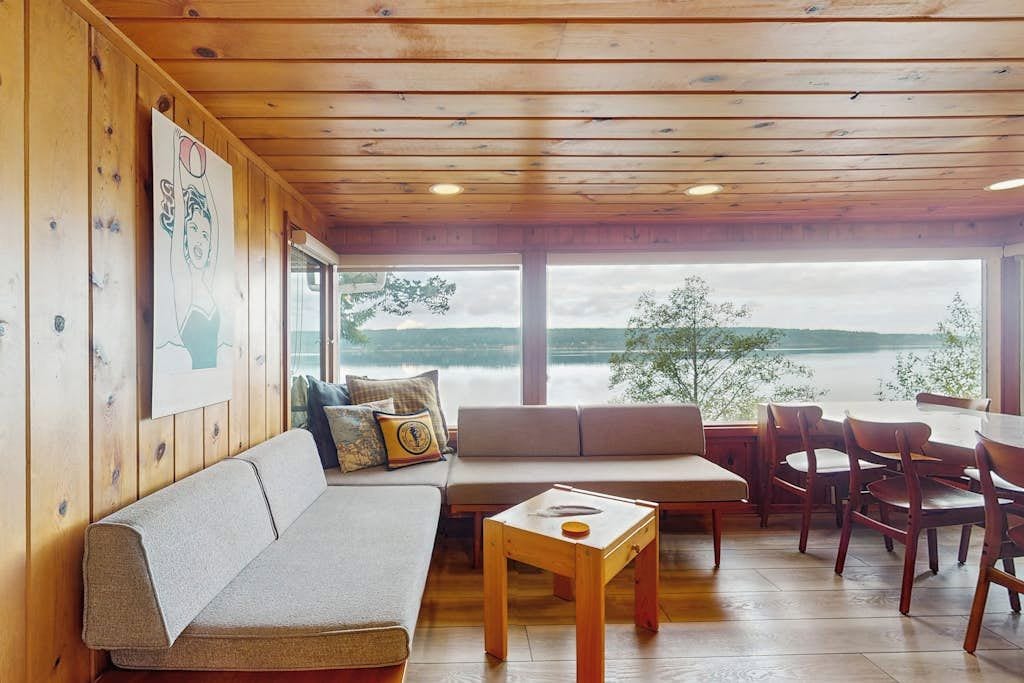 A wooden interior with a sofa, dining table, and chairs, overlooking a body of water.