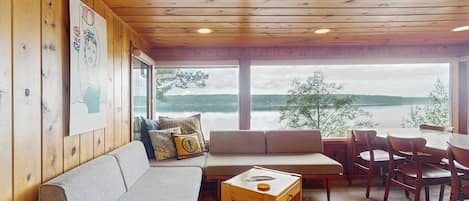 Un interior de madera con sofá, mesa de comedor y sillas, con vista a un cuerpo de agua.