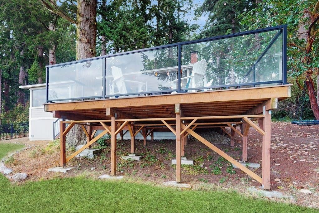A modern deck structure with a glass enclosure, supported by wooden beams, in a wooded area.