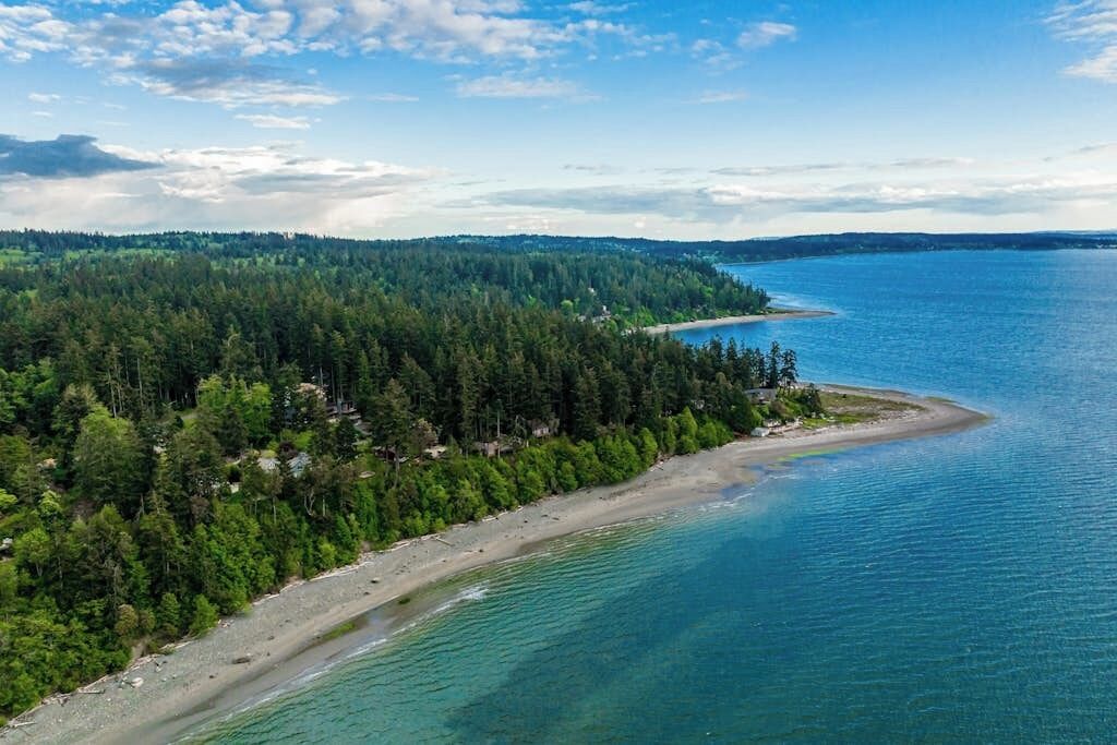 Luftaufnahme einer Küstenlandschaft mit einer bewaldeten Halbinsel und einem Strand.