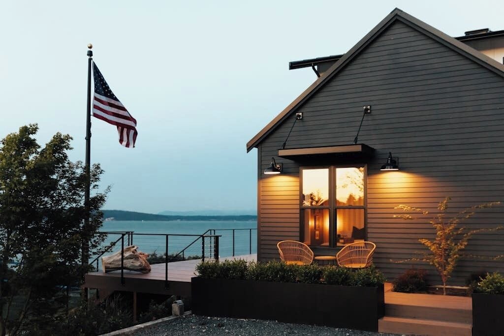 A modern house with a deck overlooking a body of water, an American flag, and two wicker chairs.