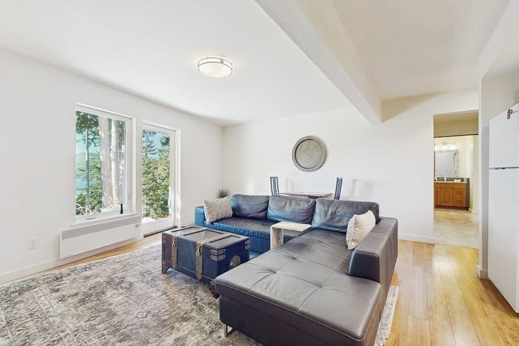 Un salon moderne avec un canapé d’angle, une table basse et un tapis. De grandes fenêtres offrent une vue sur les arbres.