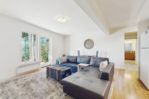 A modern living room with a sectional sofa, a coffee table, and a rug. There are large windows with a view of trees. - Cheerful Orcas Island Forest Getaway with Stunning Mountain and Sea Views (Eastsound)