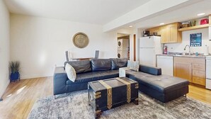 A modern living room with a sectional sofa, a coffee table, and a rug. There is a kitchen area with cabinets and appliances.