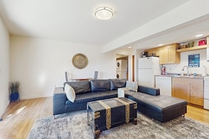 A modern living room with a sectional sofa, a coffee table, and a rug. There is a kitchen area with cabinets and appliances.