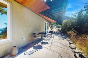 Une terrasse avec un sol en pierre, une table et des chaises, et une grande fenêtre donnant sur un paysage.
