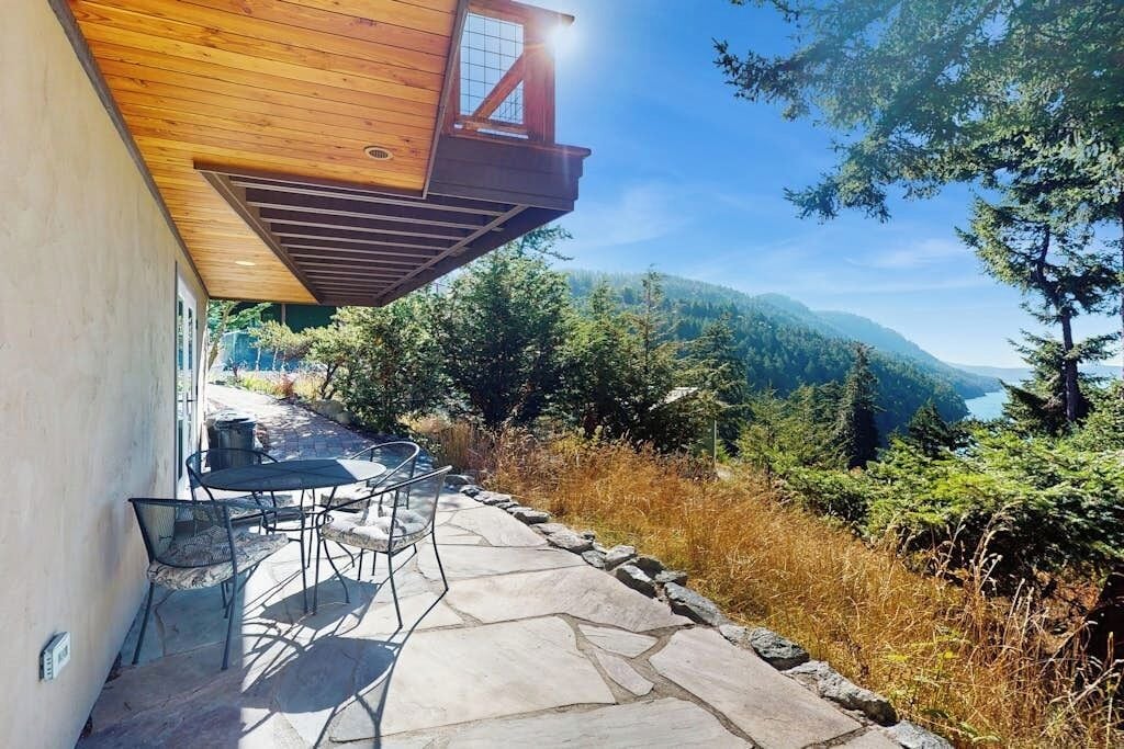 Une terrasse en pierre avec mobilier d’extérieur, une terrasse en bois et une vue sur une chaîne de montagnes boisée.