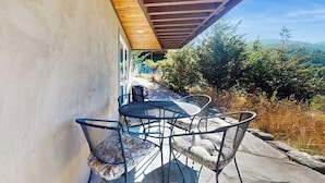 Een terras buiten met een tafel en stoelen, een houten plafond en uitzicht op bomen en heuvels.