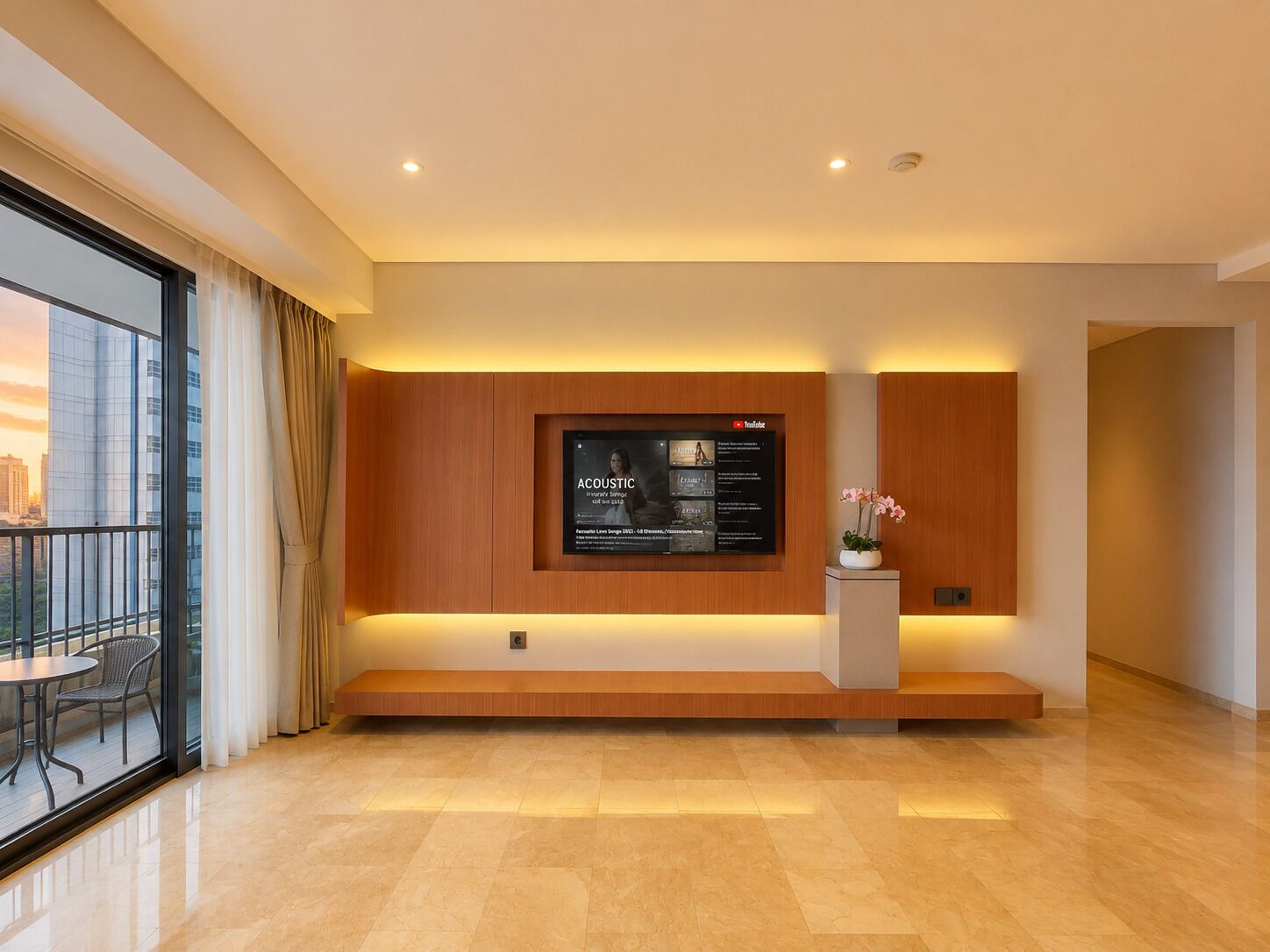 A modern living room with a flat-screen TV mounted on a wooden panel wall, a small white table with a potted plant, and a glass door leading to an outdoor balcony.